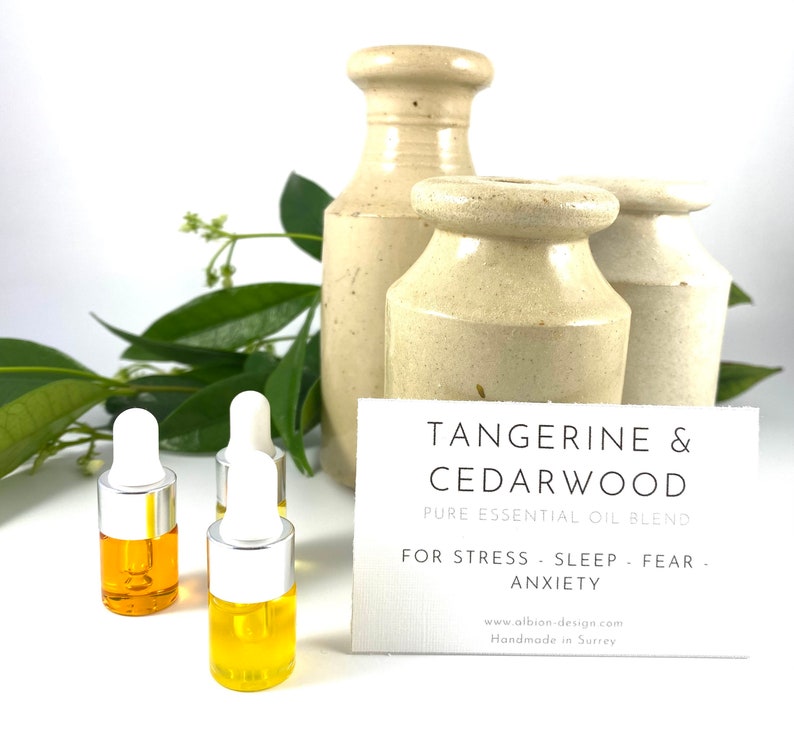 May include: Three small amber glass bottles with silver droppers filled with a yellow liquid. The bottles are on a white surface in front of a card that reads "Tangerine & Cedarwood Pure Essential Oil Blend For Stress - Sleep - Fear - Anxiety www.albion-design.com Handmade in Surrey".