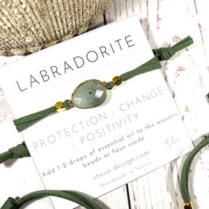 May include: Green suede bracelet with a labradorite gemstone and gold beads. The bracelet is tied with a knot and has a tag that reads "Labradorite Jewelry, Protection - Change - Positivity, Add 1-2 drops of essential oil to the wooden beads or faux suede, stosa-design.com, Handmade in Surrey".