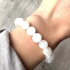 Puede incluir: Pulsera de cuentas blancas. La pulsera presenta cuentas blancas grandes y redondas ensartadas. Las cuentas tienen una superficie ligeramente texturizada. La pulsera se lleva en la muñeca.