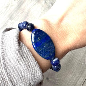 Könnte beinhalten: Ein dunkelblaues Perlenarmband mit einem großen, ovalen Lapislazuli-Stein. Das Armband besteht aus kleineren, runden Perlen und einem polierten Mittelstein. Das Armband wird am Handgelenk getragen und zeigt die Farbe und Textur der Edelsteine.