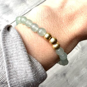 Puede incluir: Un brazalete hecho de cuentas de color verde claro con detalles dorados. El brazalete se lleva en la muñeca.