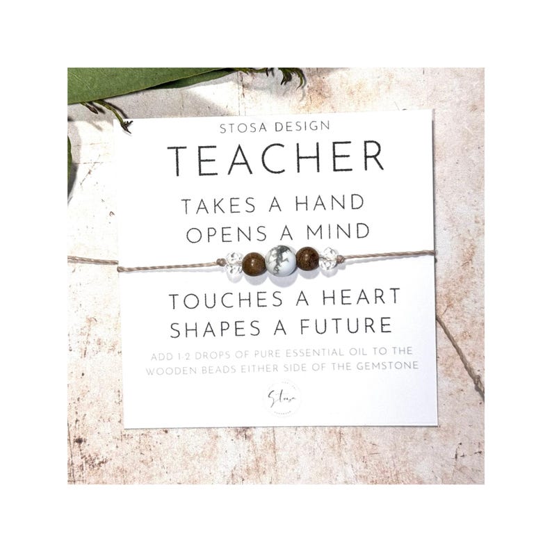 Puede incluir: Una tarjeta blanca con texto marr&oacute;n que dice "STOSA DESIGN TEACHER TAKES A HAND OPENS A MIND TOUCHES A HEART SHAPES A FUTURE ADD 1.2 DROPS OF PURE ESSENTIAL OIL TO THE WOODEN BEADS EITHER SIDE OF THE GEMSTONE Slar". Una pulsera de cuentas marrones y blancas est&aacute; en la tarjeta.