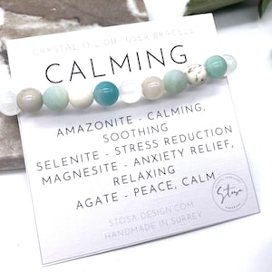 Puede incluir: Un brazalete difusor de aceite de cristal con una tarjeta blanca que dice "CALMING" y enumera las piedras y sus propiedades: Amazonita - calmante, relajante; Selenita - reducción del estrés; Magnesita - alivio de la ansiedad, relajante; Ágata - paz, calma. La tarjeta también incluye el nombre de la empresa "Stoya" y la dirección del sitio web "stosa-design.com".