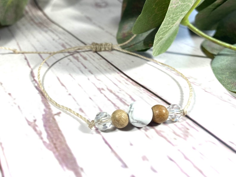 Puede incluir: Un brazalete sencillo hecho con materiales naturales. Tiene cuentas blancas y marrones, cuentas de cristal transparentes y un cord&oacute;n beige anudado.