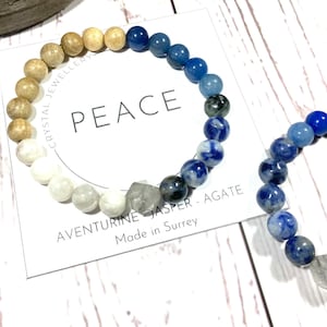 以下が含まれることがあります： 白いカードに「PEACE」と書かれた、青と茶色のビーズのブレスレット。カードには「AVENTURINE - JASPER - AGATE」と「Made in Surrey」の文字も書かれている。