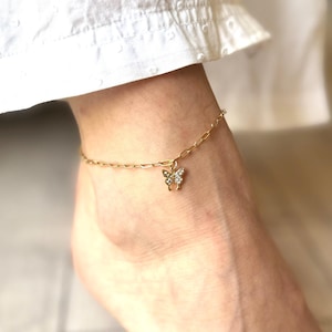 May include: Gold-coloured anklet featuring a butterfly charm. The butterfly is embellished with small, sparkling gemstones. The anklet is worn on an ankle, with white fabric visible in the background.
