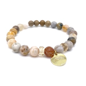 Può includere: Un braccialetto con perline e un ciondolo a forma di arcobaleno. Il braccialetto è realizzato con perline di diverse tonalità di marrone, beige e bianco.