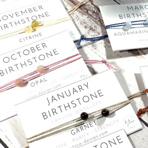 May include: A set of colourful string bracelets with birthstones for each month of the year. The bracelets are displayed on white cards with the month and birthstone name printed on them. The cards also include the words "Crystal Jewellery" and "stosa-design.com".