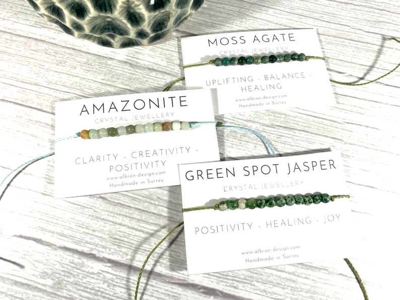 Op de afbeelding: Drie armbanden gemaakt van natuurlijke stenen en touw. De armbanden zijn gelabeld met het type steen en de bijbehorende helende eigenschappen. De stenen zijn Amazoniet, Mossaгат en Groene Vlekjaspis.