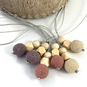 May include: A necklace with a variety of beads, including purple, pink, and beige lava rock beads, wooden beads, and beige round beads. The necklace is made with a gray cord.