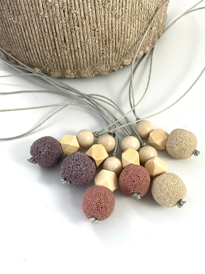 May include: A necklace with a variety of beads, including purple, pink, and beige lava rock beads, wooden beads, and beige round beads. The necklace is made with a gray cord.
