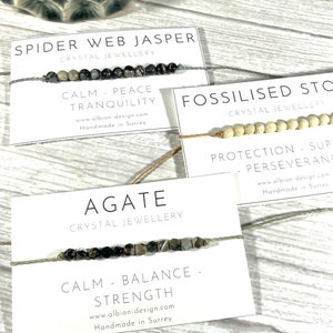 Op de afbeelding: Drie armbanden met kaartjes die hun eigenschappen beschrijven. De eerste armband is gemaakt van zwarte en grijze stenen en zegt "Spider Web Jasper Crystal Jewellery Calm - Peace - Tranquility www.albion-design.com Handmade in Surrey". De tweede armband is gemaakt van witte kralen en zegt "Fossilised Stone Crystal Jewellery Protection - Support - Perseverance www.albion design cam Handmade in Surtes". De derde armband is gemaakt van zwarte en grijze stenen en zegt "Agate Crystal Jewellery Calm - Balance - Strength www.albion-design.com Handmade in Surrey".