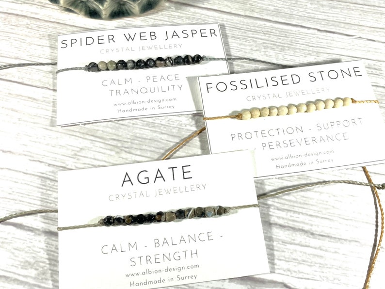 Op de afbeelding: Drie armbanden met kaartjes die hun eigenschappen beschrijven. De eerste armband is gemaakt van zwarte en grijze stenen en zegt "Spider Web Jasper Crystal Jewellery Calm - Peace - Tranquility www.albion-design.com Handmade in Surrey". De tweede armband is gemaakt van witte kralen en zegt "Fossilised Stone Crystal Jewellery Protection - Support - Perseverance www.albion design cam Handmade in Surtes". De derde armband is gemaakt van zwarte en grijze stenen en zegt "Agate Crystal Jewellery Calm - Balance - Strength www.albion-design.com Handmade in Surrey".
