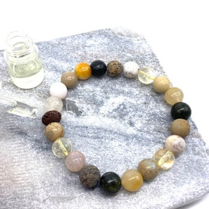 May include: A bracelet made of various natural stones, including white, brown, yellow, black, and green stones. The bracelet is on a light gray surface with a small clear glass bottle with a dropper.