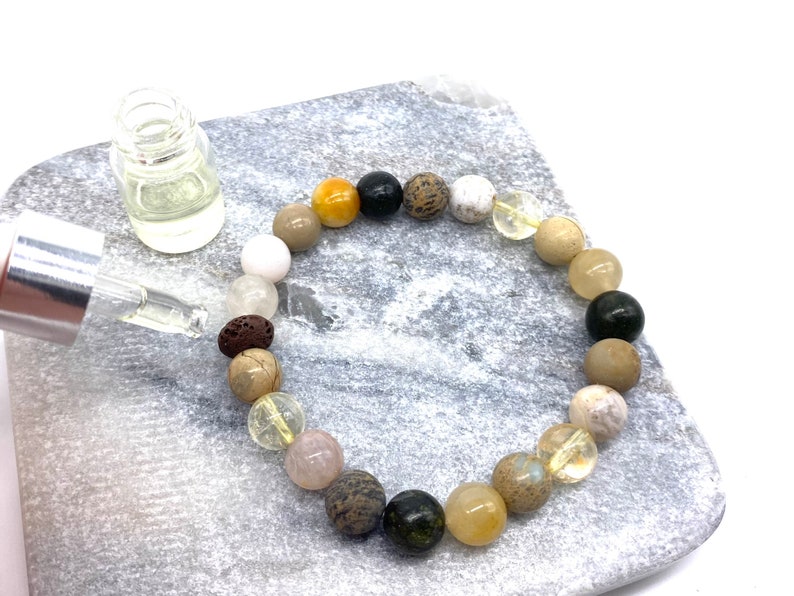 May include: A bracelet made of various natural stones, including white, brown, yellow, black, and green stones. The bracelet is on a light gray surface with a small clear glass bottle with a dropper.