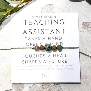 May include: A green and brown beaded bracelet with the text "STOSA DESIGN TEACHING ASSISTANT TAKES A HAND OPENS A MIND TOUCHES A HEART SHAPES A FUTURE ADD 1 2 DROPS OF PURE ESSENTIAL OIL TO THE WOODEN BEADS EITHER SIDE OF THE GEMSTONE St"
