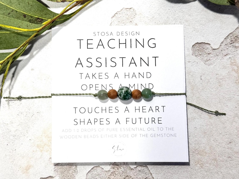 May include: A green and brown beaded bracelet with the text "STOSA DESIGN TEACHING ASSISTANT TAKES A HAND OPENS A MIND TOUCHES A HEART SHAPES A FUTURE ADD 1 2 DROPS OF PURE ESSENTIAL OIL TO THE WOODEN BEADS EITHER SIDE OF THE GEMSTONE St"