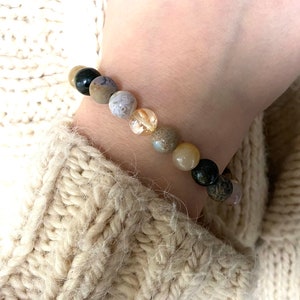 May include: A bracelet made of natural stones in shades of brown, beige, and black. The bracelet has a variety of different stones, including agate, jasper, and quartz.