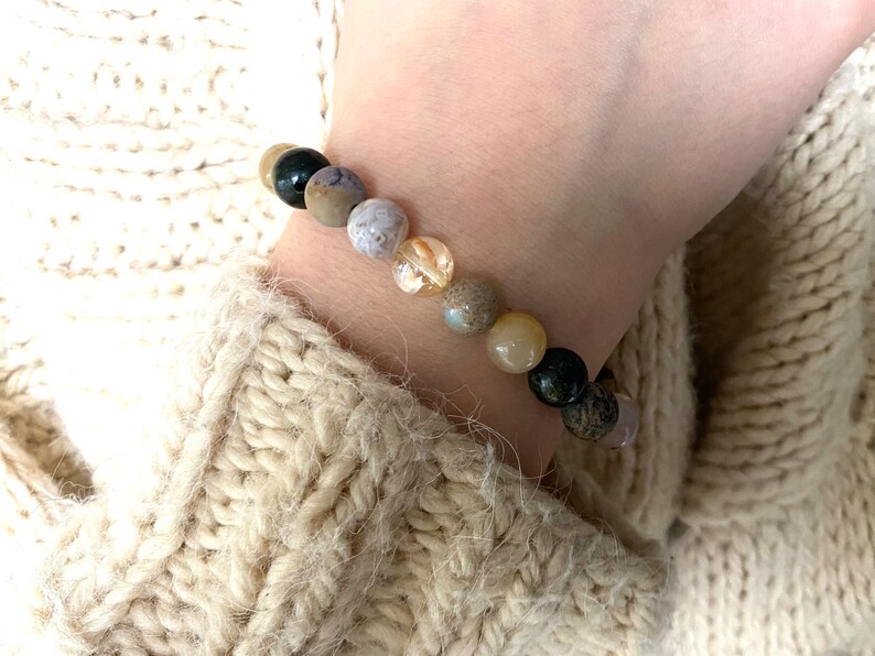 May include: A bracelet made of natural stones in shades of brown, beige, and black. The bracelet has a variety of different stones, including agate, jasper, and quartz.