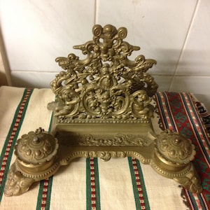 May include: Ornate antique brass inkwell with a detailed cherub and scroll design. The inkwell features two ink pots with decorative lids and a central pen rest. The piece is set on a striped fabric.