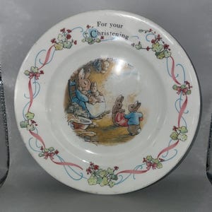 May include: A white ceramic plate with a blue and pink border. The centre of the plate features a picture of Peter Rabbit and his family. The text "For your Christening" is written on the plate.
