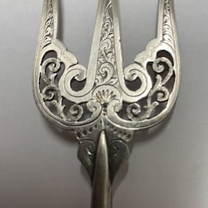 Op de afbeelding: Een zilveren vork met een decoratief, sierlijk ontwerp. De vork heeft drie tanden en een lange, slanke steel.