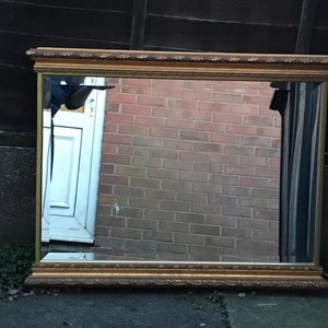 Pode incluir: Um espelho grande e retangular com uma moldura dourada ornamentada. O espelho reflete uma parede de tijolo e uma porta.