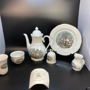 Puede incluir: Un juego de té de porcelana blanca con un diseño de escena de invierno. El juego incluye una tetera, una jarra de crema, un azucarero, una taza y una pequeña campana. El juego está decorado con ribetes dorados y presenta el texto "Colonial Haus of Germany" y "Quality Porcelain".