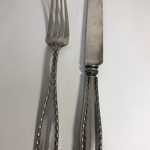 Puede incluir: Un tenedor y un cuchillo de plata con mangos ornamentados. El tenedor tiene un borde decorativo y la letra "D" grabada en el mango. El cuchillo tiene un borde decorativo y un mango retorcido.