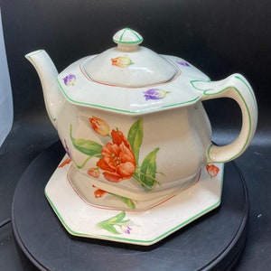 May include: A white teapot with a green trim and floral design featuring orange and purple flowers. The teapot has a matching saucer with a similar floral design.