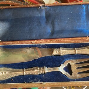 Puede incluir: Un juego de servicio de plata con un diseño ornamentado, que incluye un cuchillo para mantequilla y un tenedor con abridor de botellas, en una caja forrada de terciopelo azul.