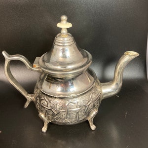 May include: Silver teapot with an ornate design and a white knob on the lid.