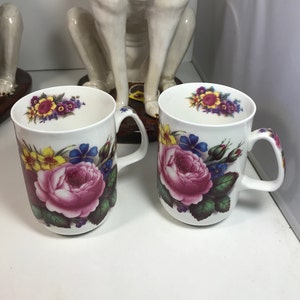 May include: Two white ceramic mugs with floral designs. The mugs feature a large pink rose with yellow, blue, and purple flowers surrounding it. The mugs have a white background and a black rim.