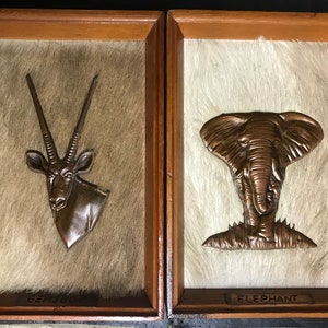 May include: Two framed wall decor pieces with a faux fur background. The left piece features a bronze-colored oryx head with long horns. The right piece features a bronze-colored elephant head. The left piece is labeled "Gemsbok" and the right piece is labeled "Elephant".