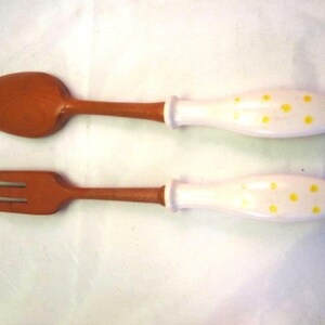 Puede incluir: Una cuchara de madera y un tenedor de madera, cada uno con un acabado marrón liso en la cabeza y los dientes. Los mangos son de material blanco decorado con pequeños puntos amarillos, creando un contraste entre la madera y el mango.
