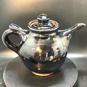 May include: A glossy black ceramic teapot featuring a rounded body, a curved handle, and a tapered spout. The surface is decorated with small, scattered orange polka dots. A matching lid with a small knob sits securely on the top opening.