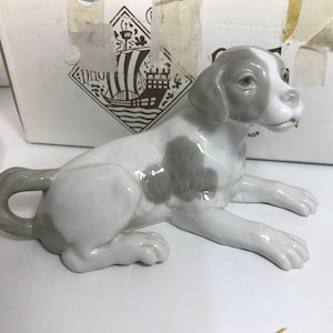 Puede incluir: Una figurita de porcelana blanca de un perro acostado. El perro tiene marcas grises en su cuerpo y cabeza. La figurita está en una caja con un logotipo de un barco.