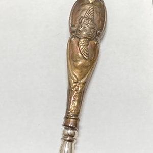 May include: A silver-toned shoe horn with an ornate handle featuring an embossed cherub design. The handle is shaped like an elongated oval and has a detailed design.