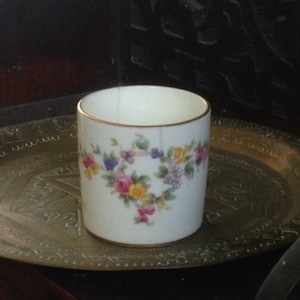 May include: A white porcelain cup with a gold rim and a floral design in shades of pink, yellow, blue, and green. The cup is sitting on a brass tray.