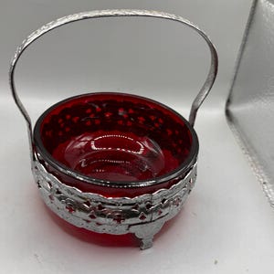Könnte beinhalten: Eine kleine, runde Schale aus tiefrotem Glas, die in einen dekorativen, silberfarbenen Metallrahmen eingesetzt ist. Der Metallrahmen weist ein kompliziertes, perforiertes Gittermuster auf und besitzt einen passenden, bogenförmigen Henkel.