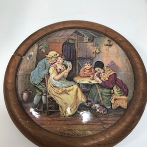 Pode incluir: Uma caixa redonda de madeira com uma tampa com dobradiças. A tampa apresenta uma ilustração colorida de quatro pessoas jogando cartas em um ambiente rústico. O texto "THE BEST CARD" está impresso na parte inferior da caixa.