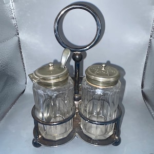 Könnte beinhalten: Ein Salz- und Pfefferstreuer-Set aus Silber und Schwarz mit einem Glasbehälter und einem Metalldeckel. Das Set steht auf einem Metallständer mit einem Ringgriff.