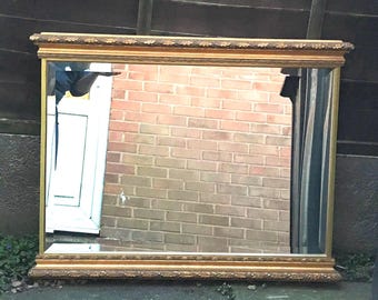 May include: A rectangular wall mirror featuring a wide, ornate gold-toned frame with detailed carvings along the top and bottom edges. The reflective glass surface is clear and set within the decorative border, providing a wide-angle view.