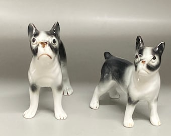 May include: Two small porcelain figurines of black and white Boston Terrier dogs. The figurines are standing on a white surface next to a British pound coin.