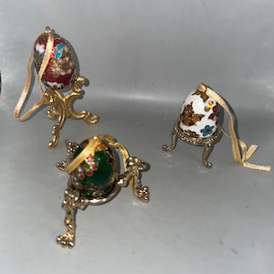 Op de afbeelding: Drie decoratieve eierornamenten met bloemmotieven en goudkleurige standaards. Eén ei is rood met blauwe en witte bloemen, een ander is wit met bruine en blauwe bloemen, en de derde is groen met rode bloemen.