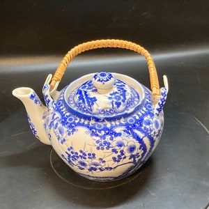 Puede incluir: Una tetera de porcelana azul y blanca con un asa de bambú. La tetera tiene un diseño floral y está decorada con pintura azul y blanca.