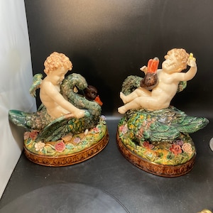 Puede incluir: Par de figuras de cerámica de querubines montando cisnes. Los cisnes son verdes, marrones y blancos con plumas detalladas. Los querubines están desnudos y tienen alas. Las bases están decoradas con flores y hojas.
