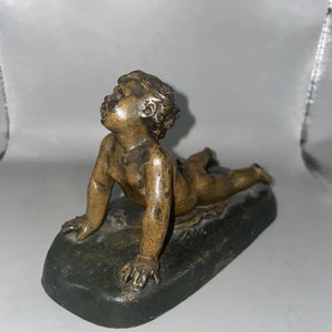 Könnte beinhalten: Eine Bronzeskulptur eines liegenden Cherubs, der nach oben schaut. Der Cherub steht auf einem dunkelgrünen Sockel.
