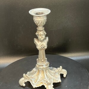 Puede incluir: Un candelero de plata con un diseño ornamentado que presenta una cabeza de querubín y una base decorativa.
