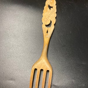 Könnte beinhalten: Holzgabel mit dekorativem Griff. Der Griff hat ein geschnitztes Design mit dem Text "1946" und "Hillebrand". Die Gabel hat vier Zinken und ist aus hellem Holz gefertigt.